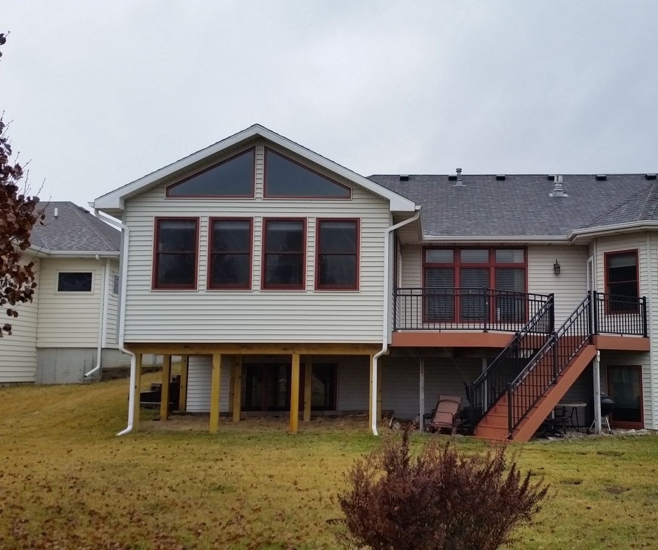 Back of a house with a sunroom, raised deck, and stairs. White siding, red window frames, and a cloudy sky.