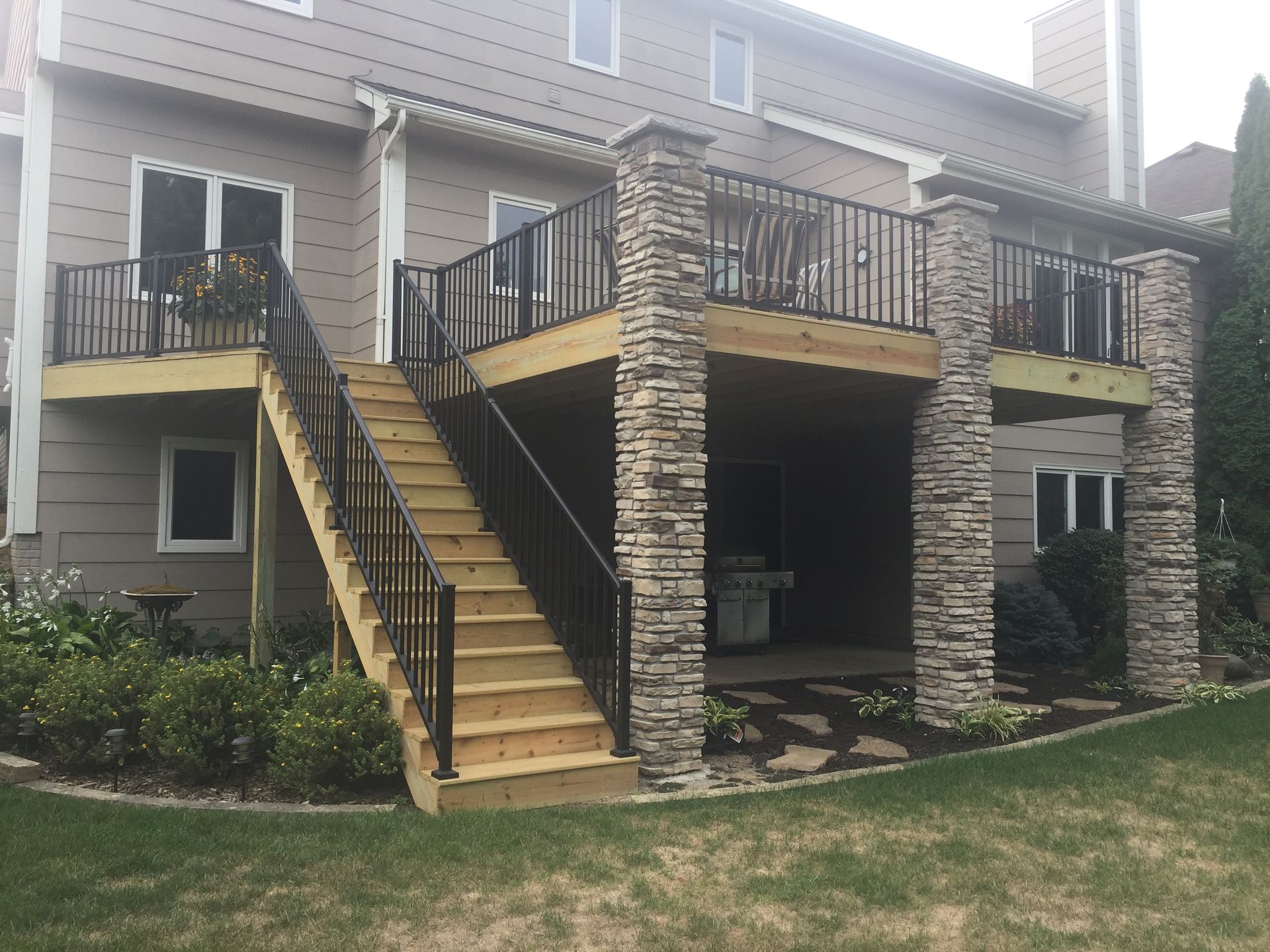 Two-story deck with stone columns, stairs, and black railings, viewed from backyard.