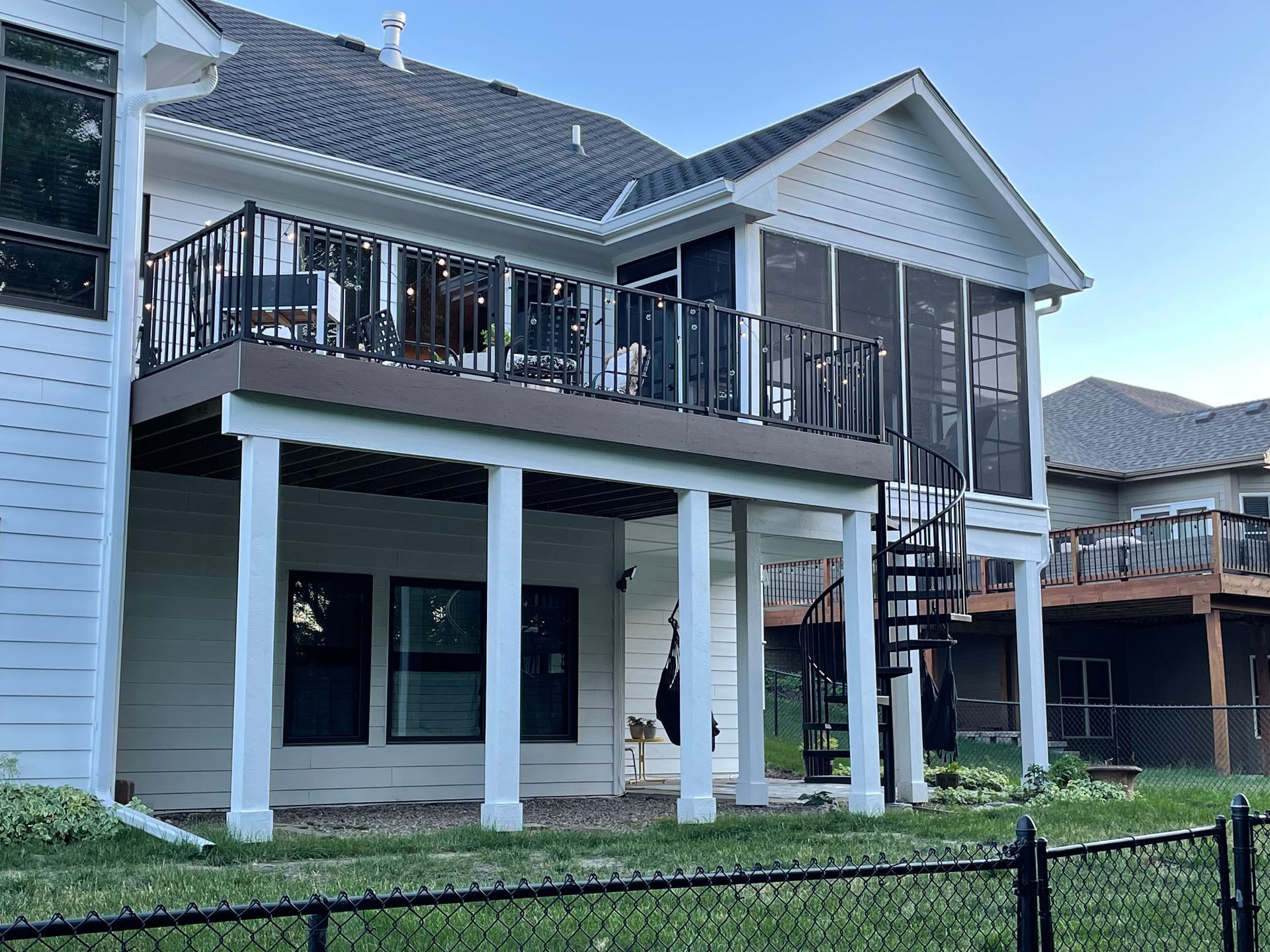 Two-story house with a deck, screened porch, and spiral staircase. Black railing, white siding, green grass.