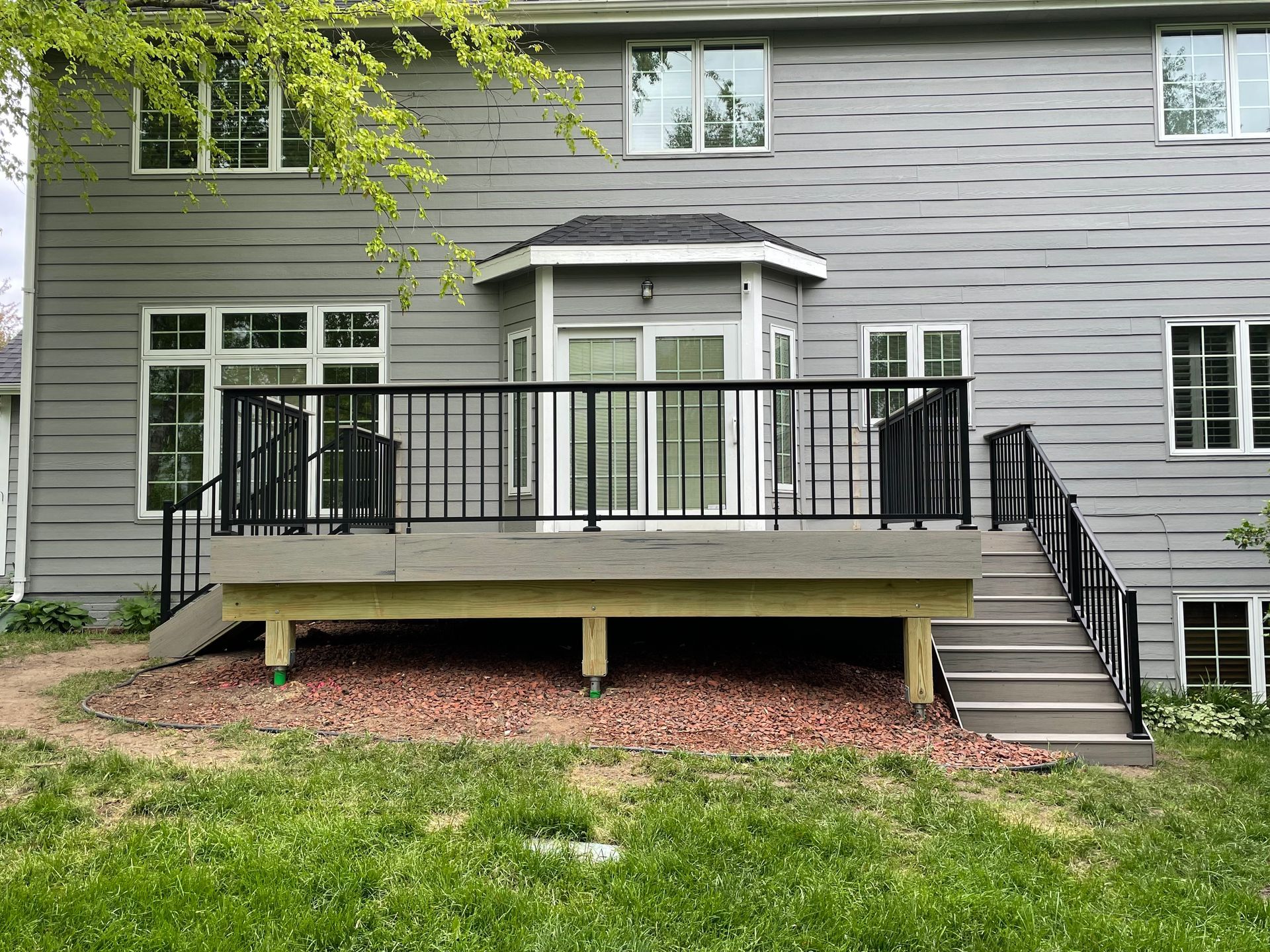 Back of a gray house with a wooden deck and black railing, steps leading down to a grassy yard.