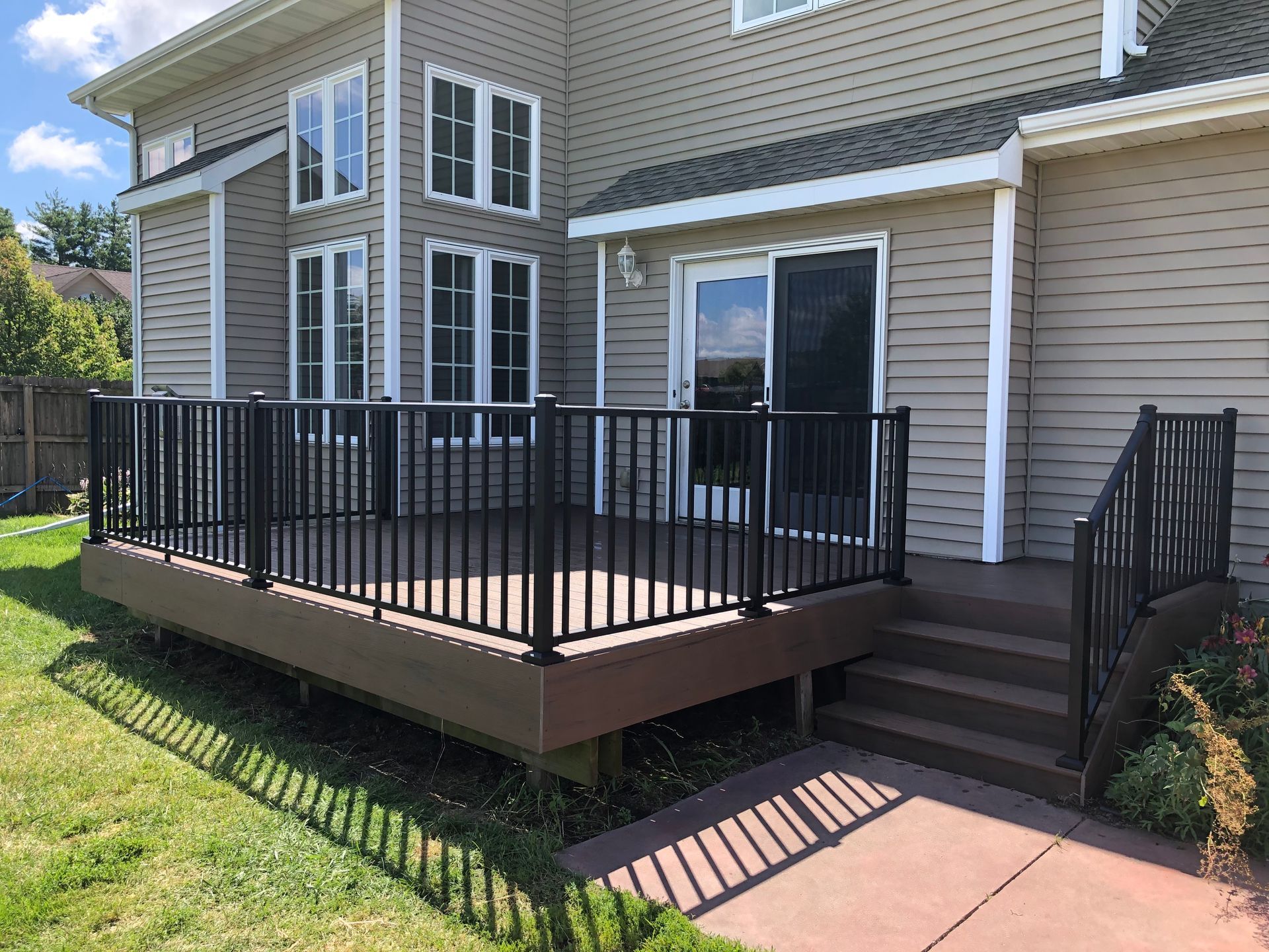 Backyard deck with black railing and steps leading to a sliding glass door. Brown decking and tan siding.