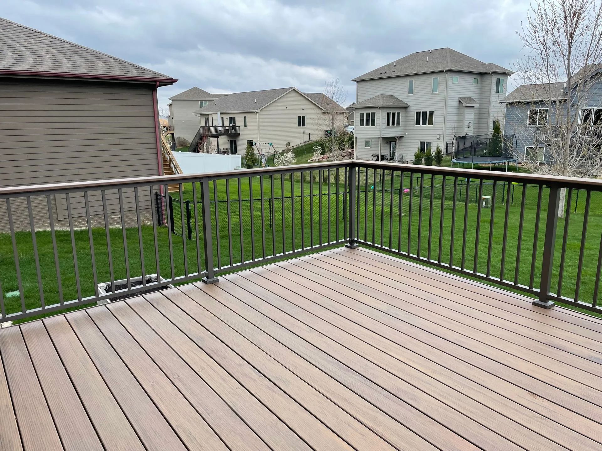 Deck with dark railing overlooking a green lawn and multiple houses on a cloudy day.