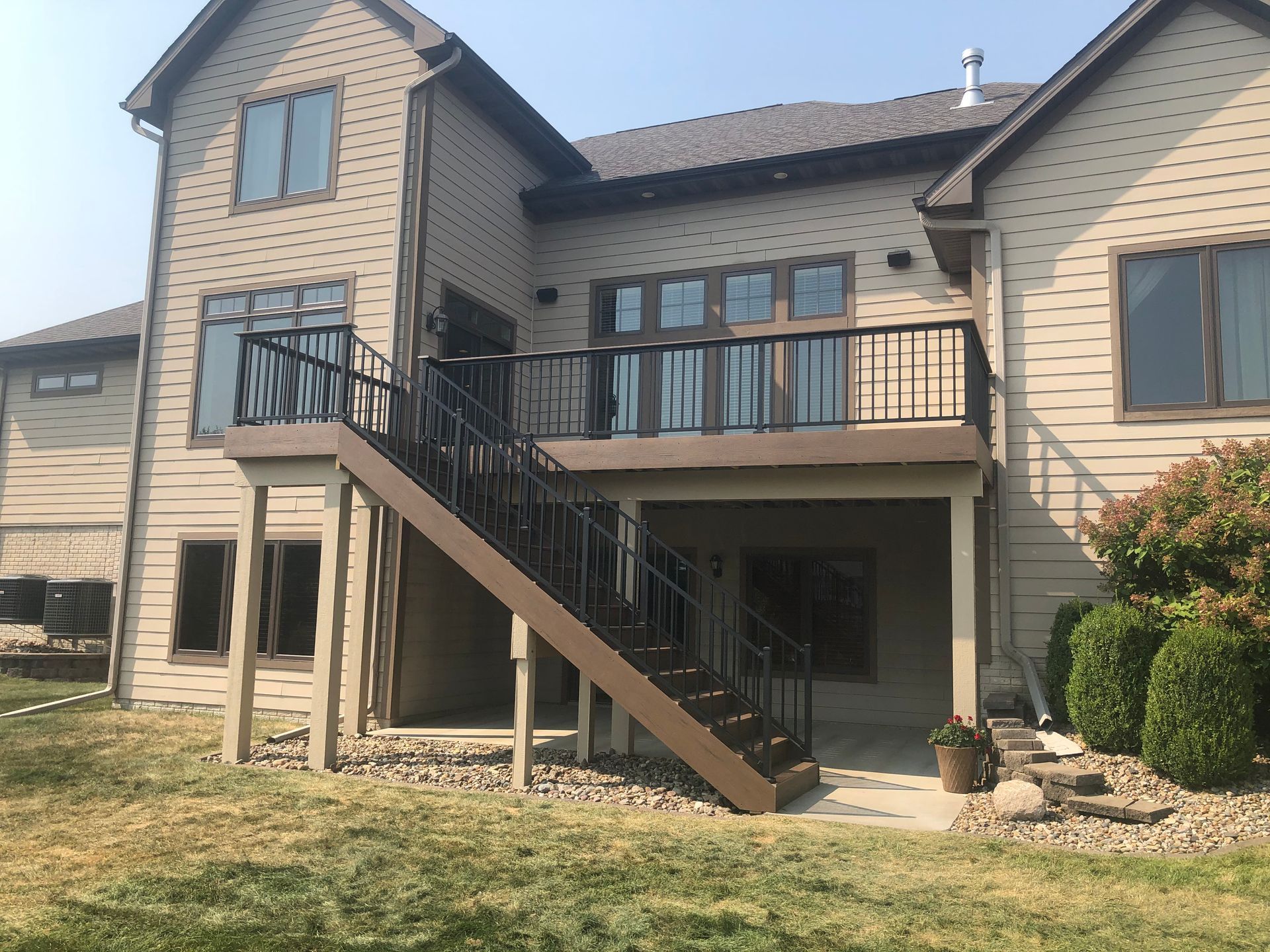 Two-story house with a deck and stairs. Brown deck and stairs, black railings, beige siding, and a grassy yard.