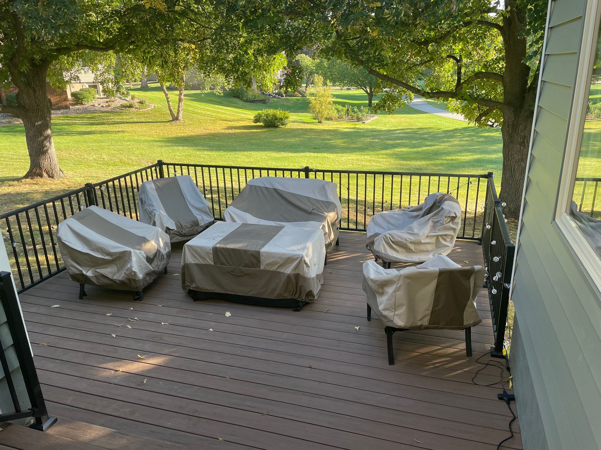 Patio furniture covered on a wooden deck, surrounded by a black railing, next to a grassy yard with trees.