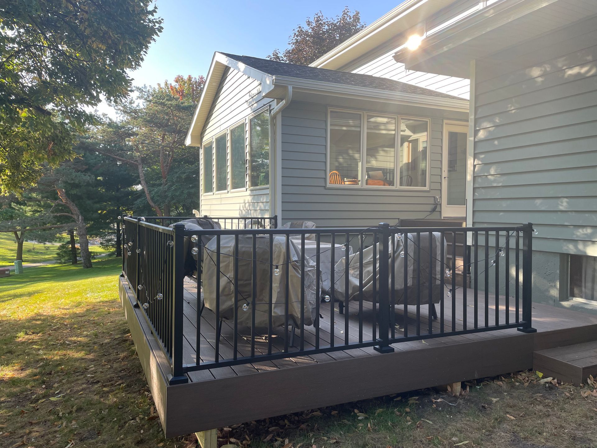 Black railing surrounds a deck with covered chairs. Attached to a gray house, overlooking a grassy yard.