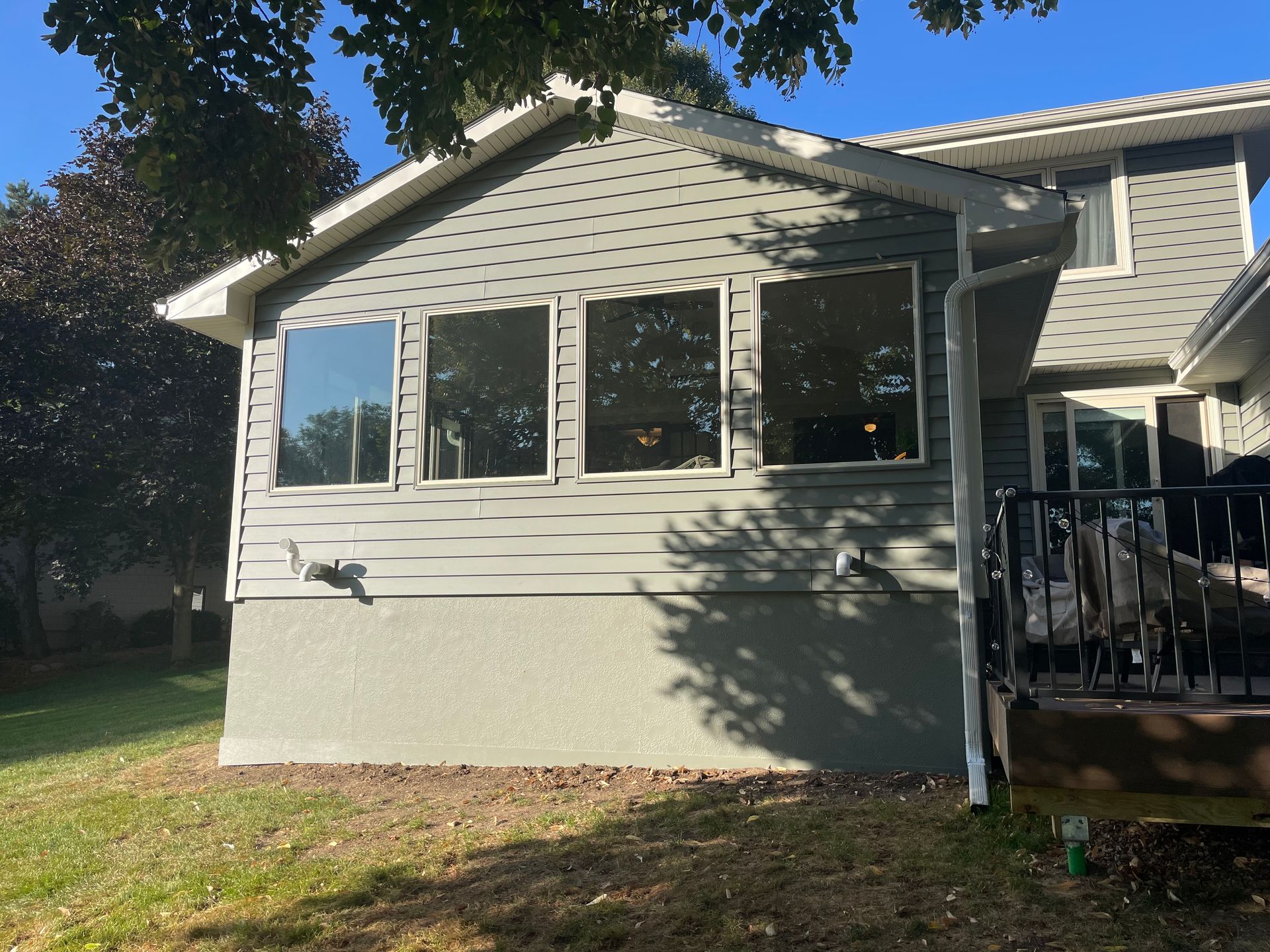 Gray house exterior with windows and stucco base. Green grass and tree in background.