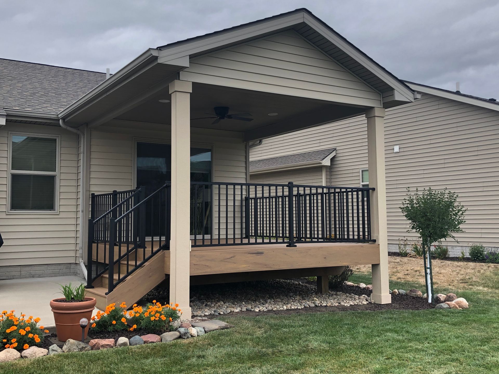 Covered deck with black railing, steps, beige siding, and a small tree in a yard.