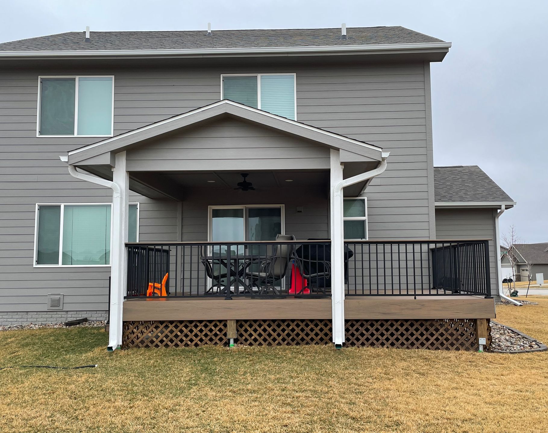Two-story house with covered deck, black railing, and lattice skirting. Gray siding, brown deck, overcast.