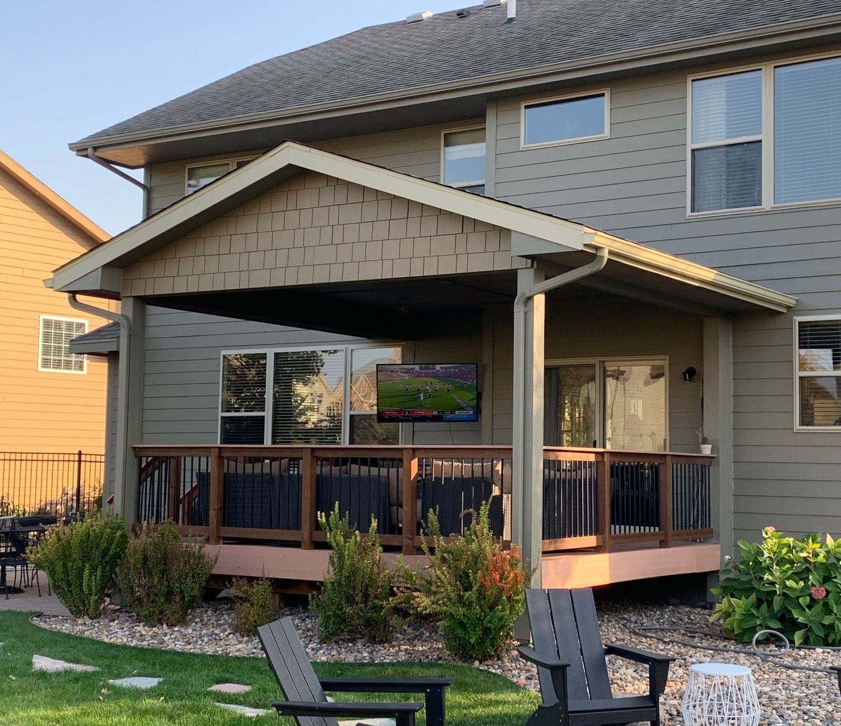 Two-story home with an outdoor deck, featuring a TV, chairs, and lush green lawn.
