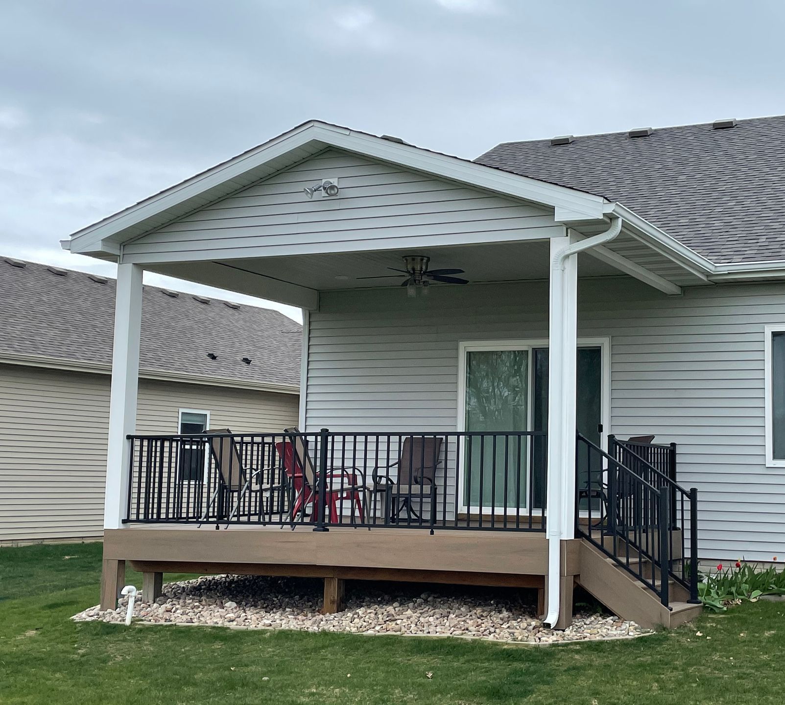 A covered wooden deck with black railing, chairs, and a sliding door. Grass and a house are in the background.