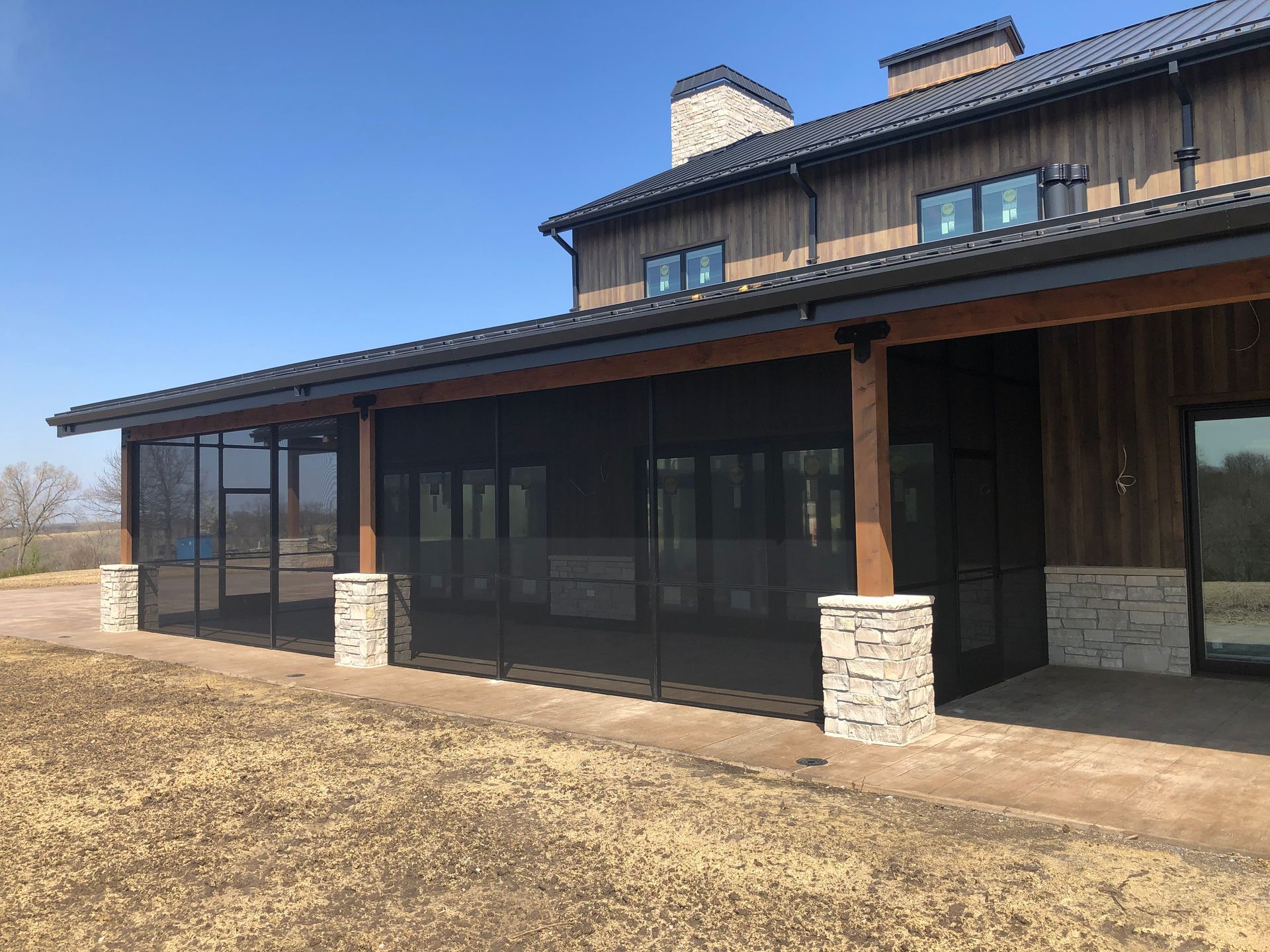 Exterior of a house with a screened-in porch supported by stone columns and wooden beams; brown siding, clear sky.