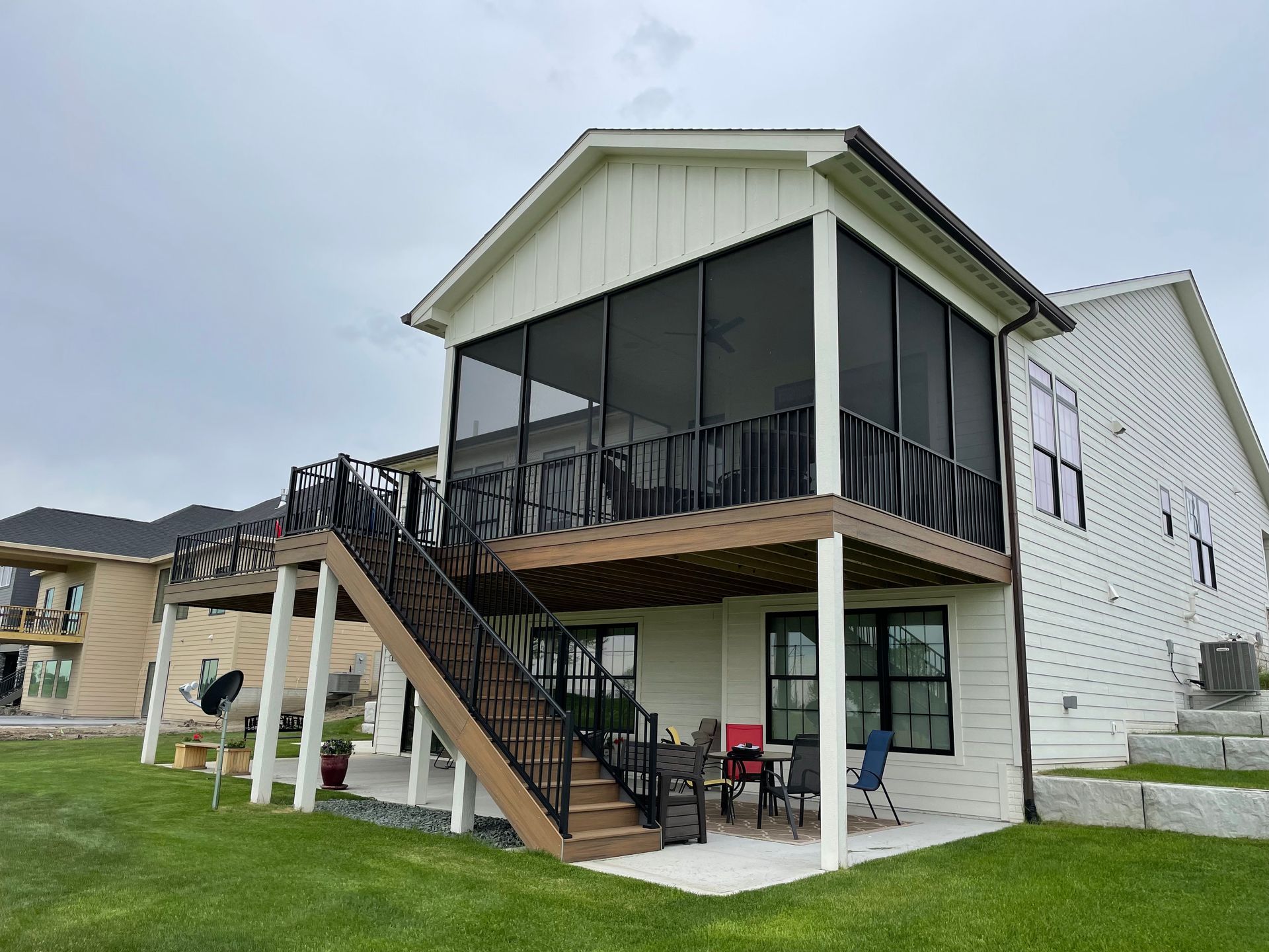 Screened porch with black railing, attached to a white house with a wooden deck, overlooking a green lawn.