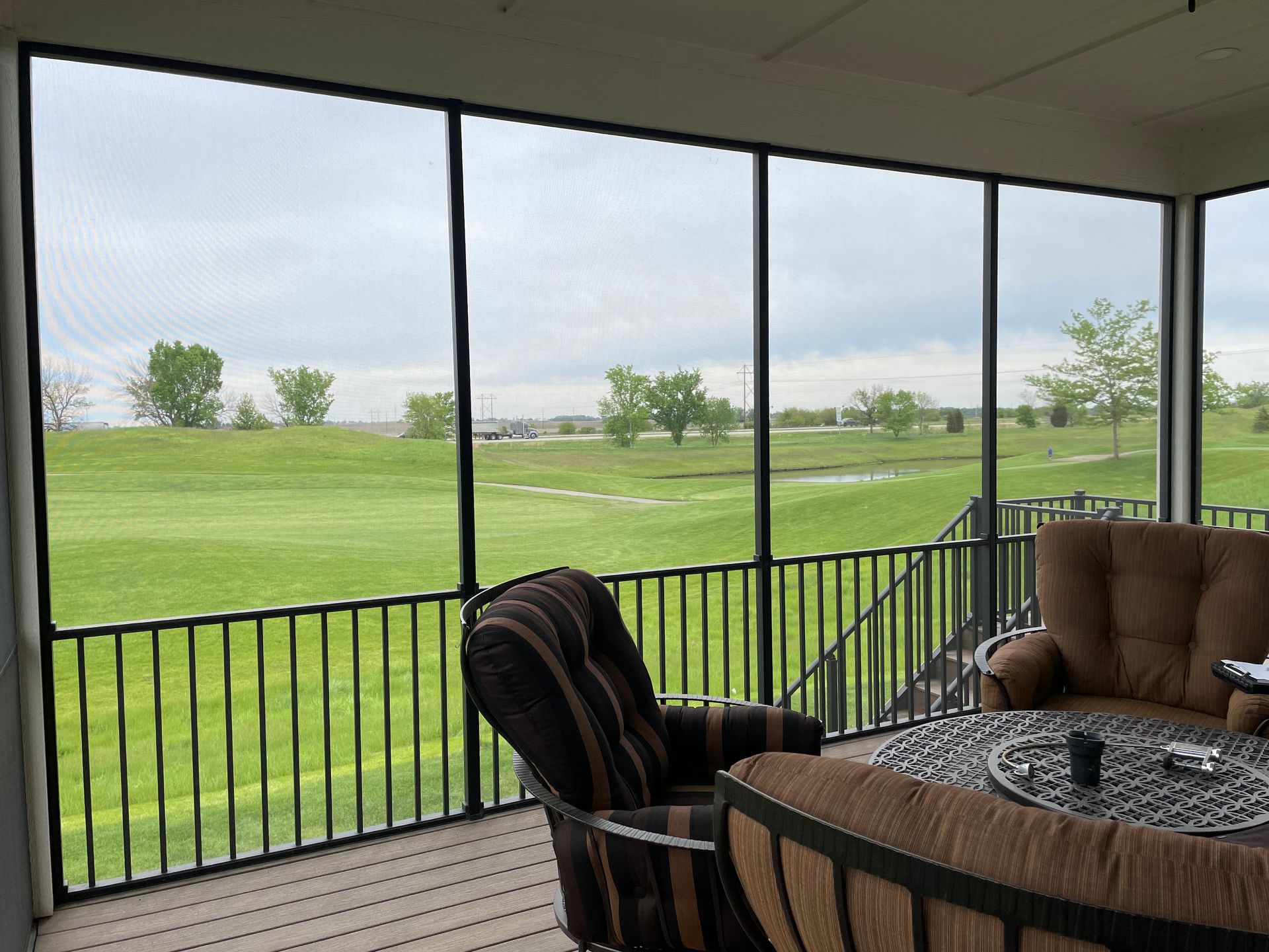 A screened porch overlooking a green landscape with trees, furniture, and a cloudy sky.