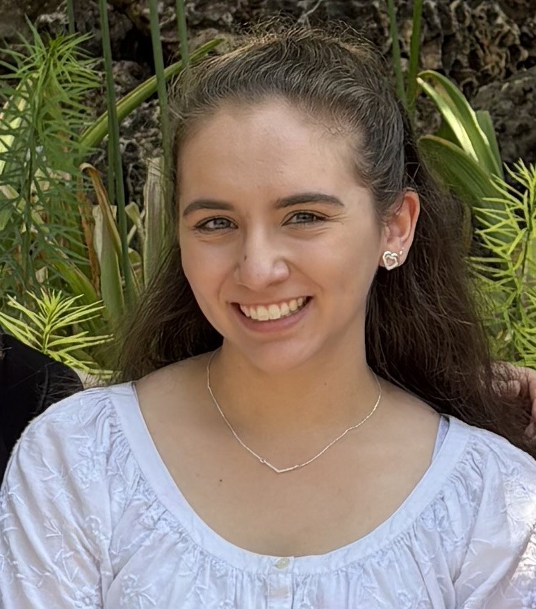 Smiling woman in a white blouse standing outdoors with green plants in the background