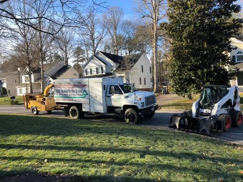 White work truck, yellow dump truck, and skid steer parked on a grassy suburban street in daylight