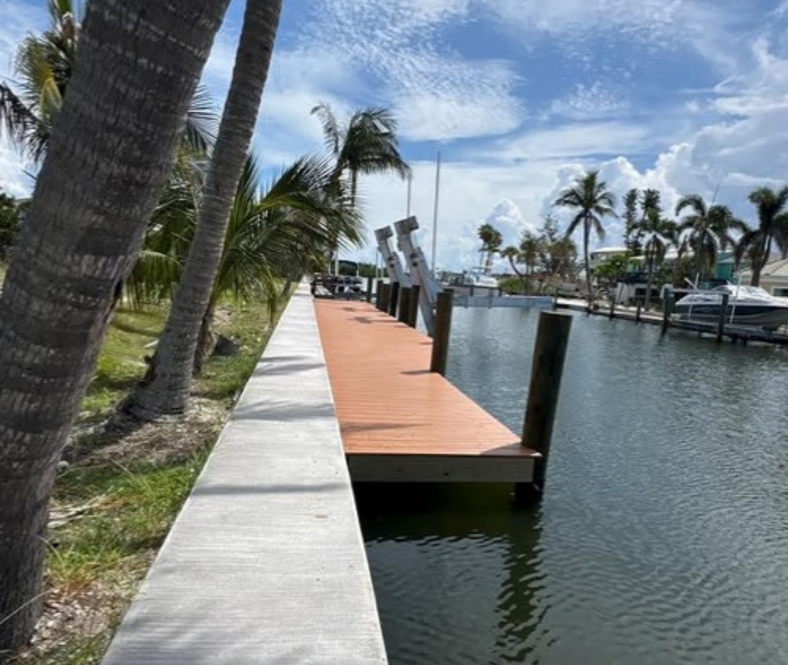 A narrow, wood-planked boat dock sits along a concrete seawall beside a calm canal, flanked by palm trees under a sunny sky.