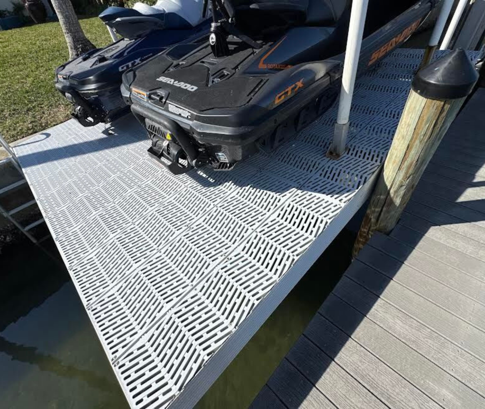 Two jet skis parked on a white, grated floating dock next to a wooden pier by a grassy shore.