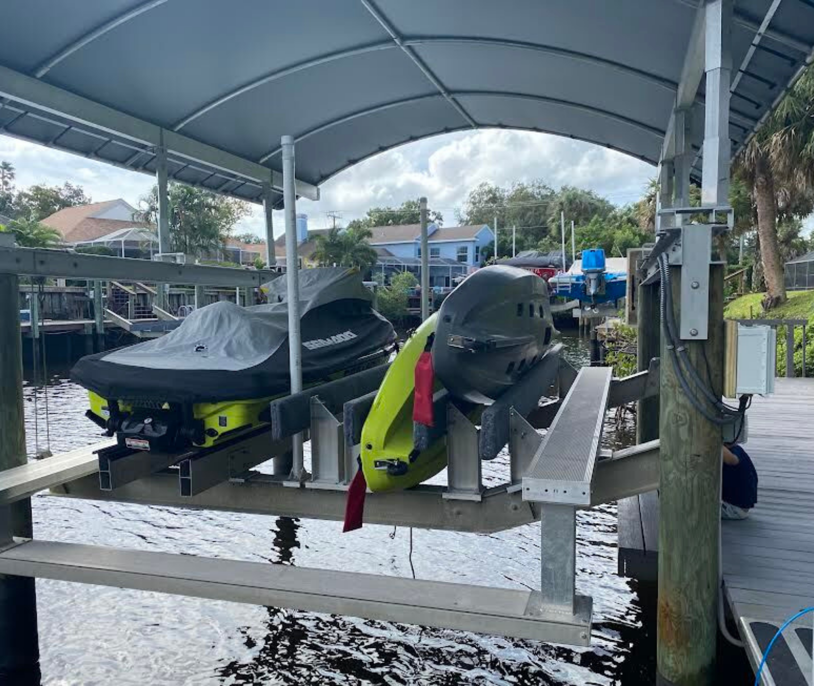 Two jet skis on a boat lift under a canopy over a canal, with a person sitting on the wooden dock to the right.