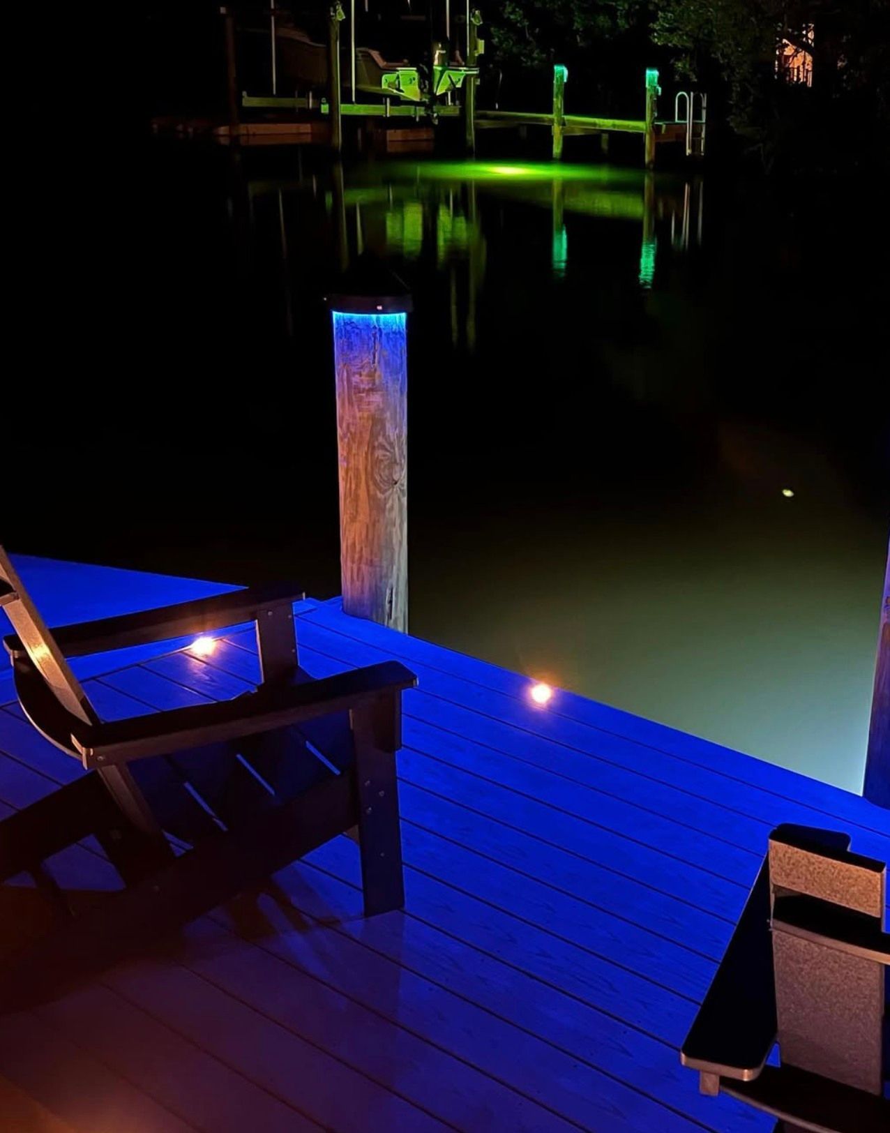 Two chairs on a wooden dock at night, illuminated by blue deck lights and green underwater dock lighting.
