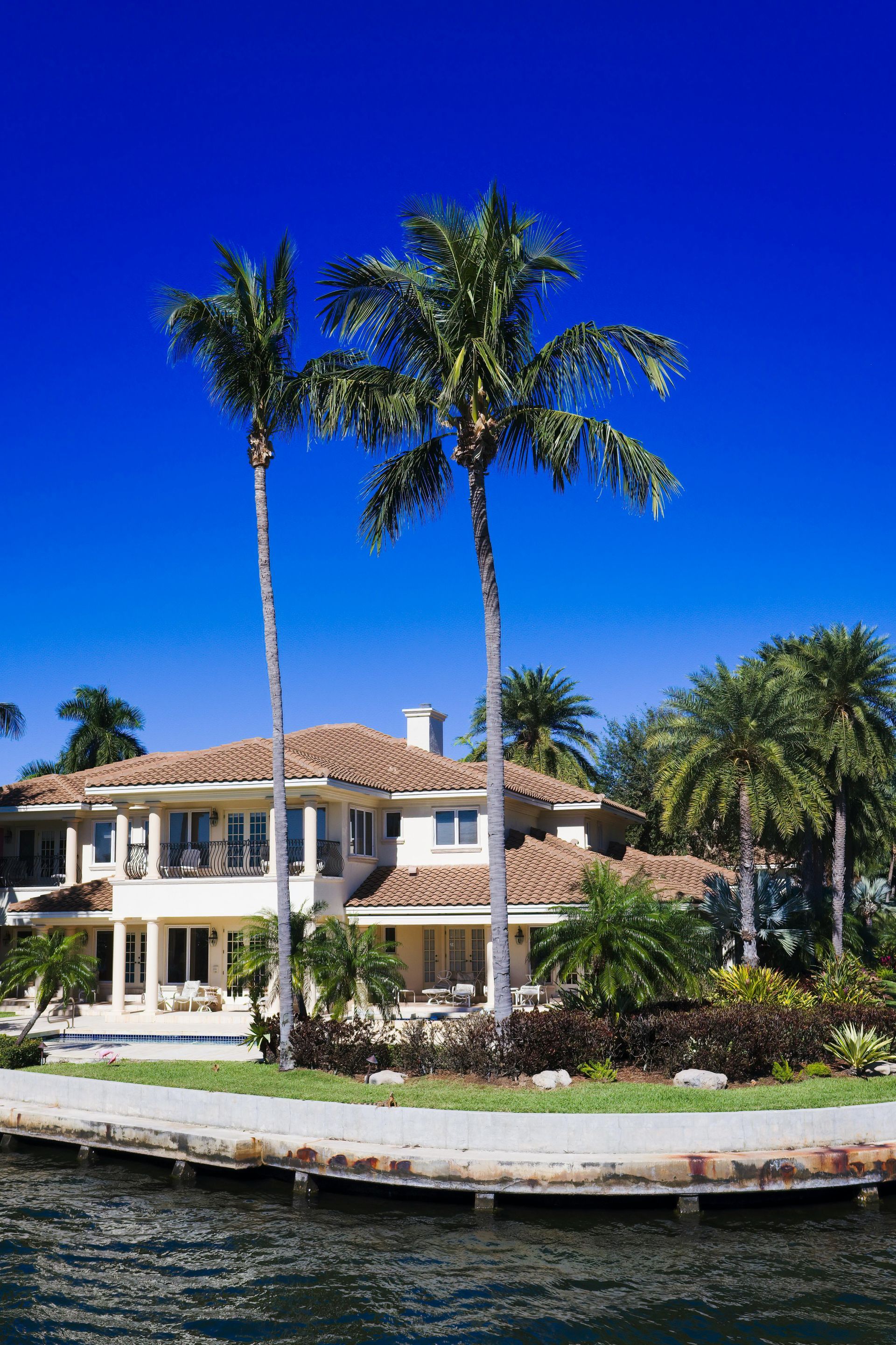 Large, light-colored Mediterranean-style home with a tile roof situated behind a canal, surrounded by palm trees.