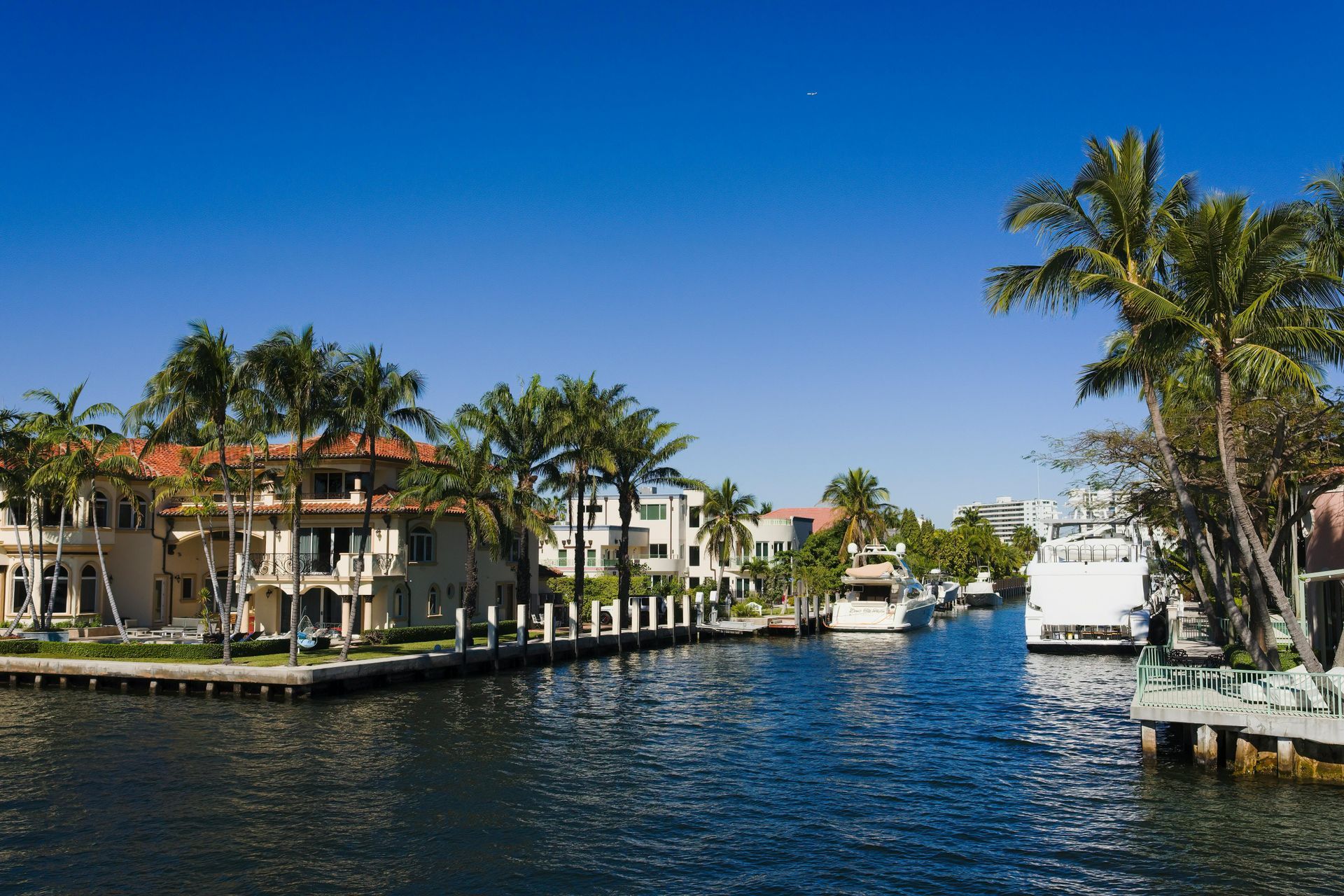 Luxury houses and palm trees line a sunny canal with docked boats under a clear blue sky.
