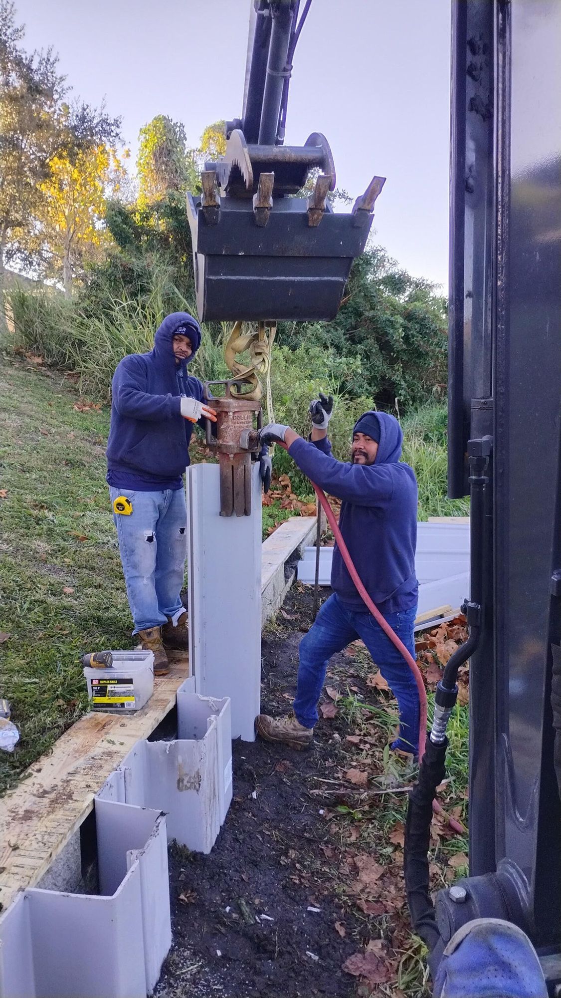 Two construction workers in blue hoodies use a mechanical bucket to install a white post at an outdoor job site.