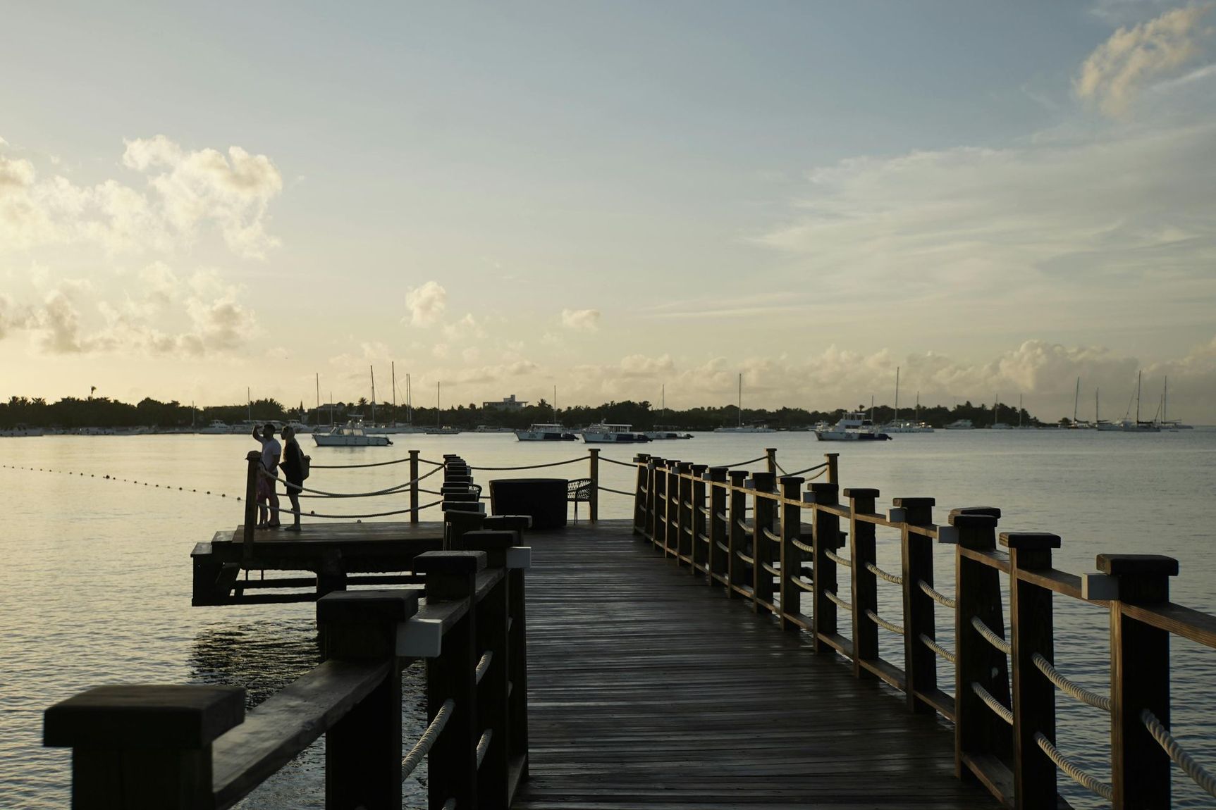Wooden pier over calm water at sunset, with distant boats and a cloudy sky
