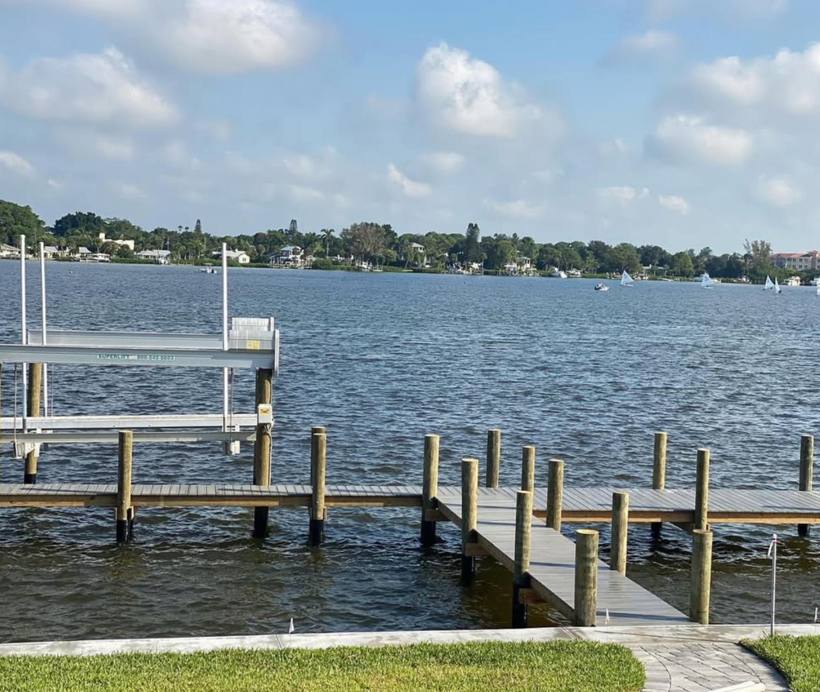 A wooden dock with a metal boat lift extends into a calm lake under a bright, partly cloudy blue sky.