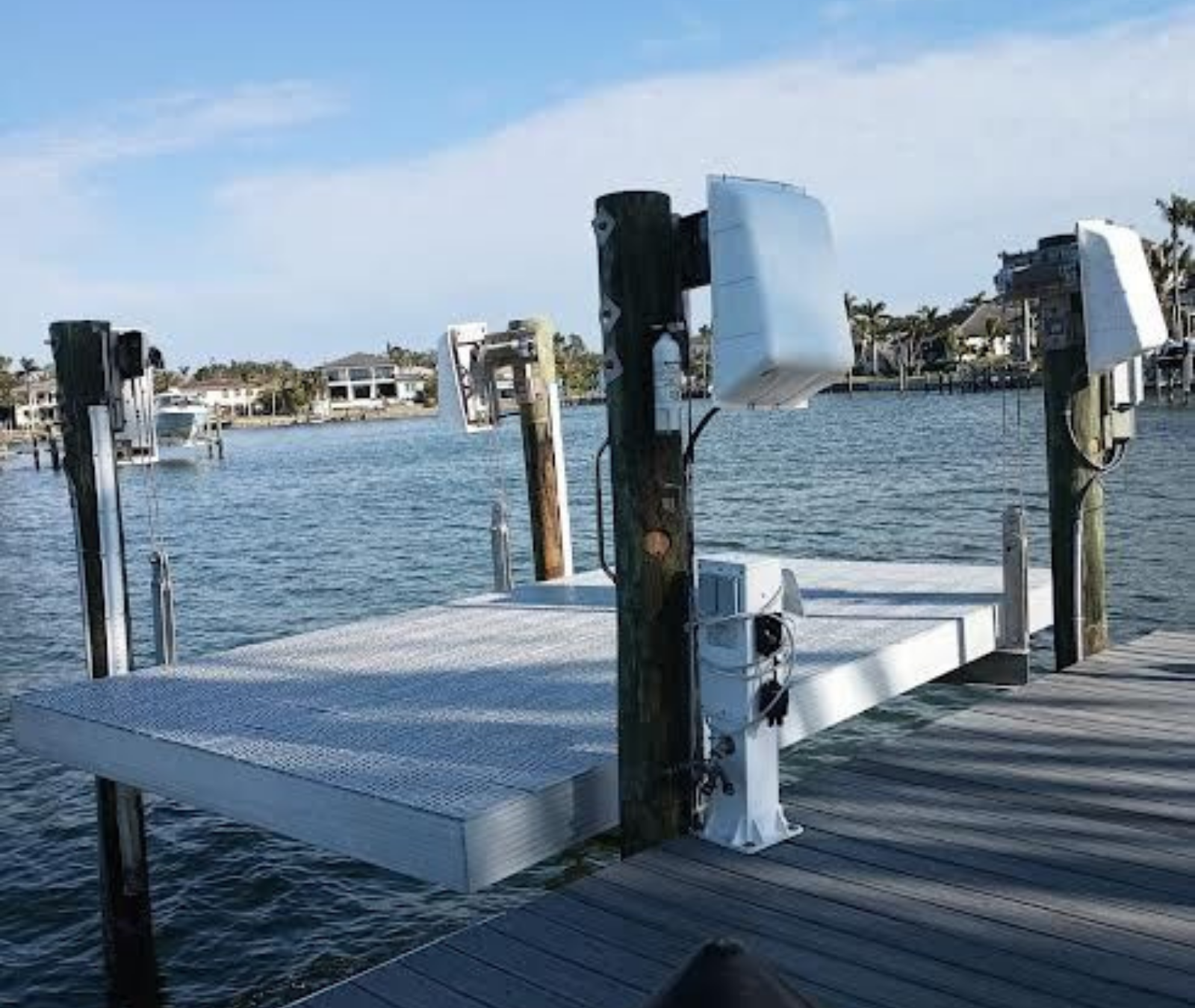 A white boat lift platform lowered into the water between four wooden pilings on a sunny day at a residential dock.
