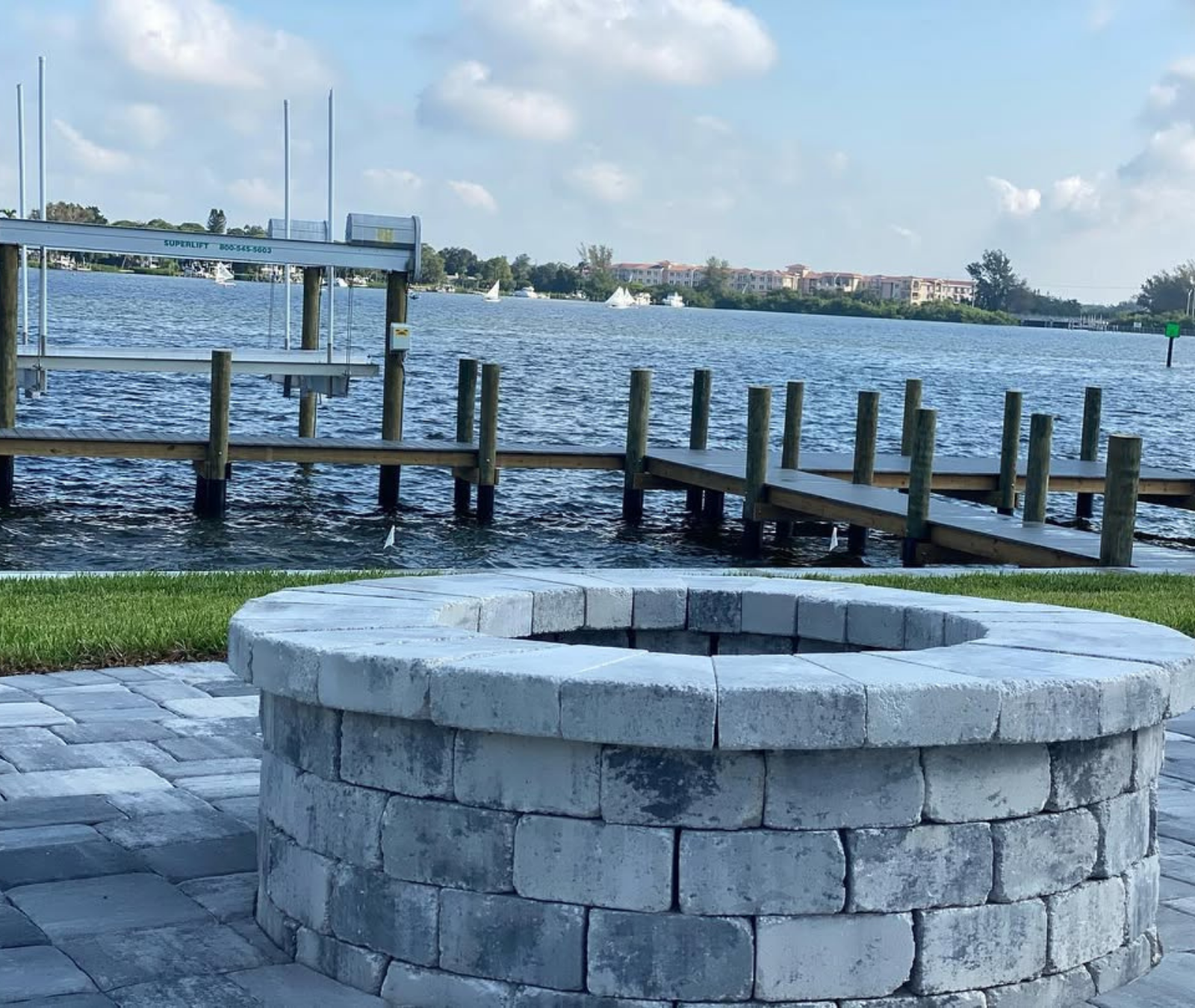 A circular gray stone fire pit on a paver patio overlooking a wooden dock and boat lift on a sunny, breezy waterfront.