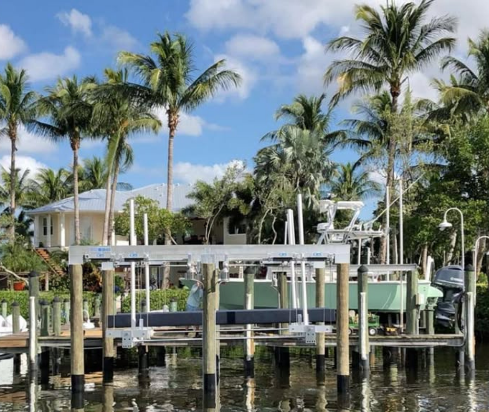 A boat lift structure stands over water in front of a house, surrounded by palm trees under a sunny blue sky.