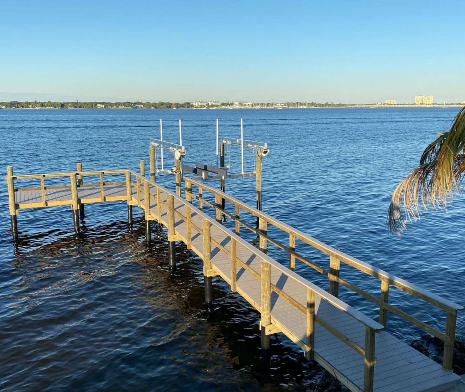 A wooden pier with railings extends into calm blue water under a clear sky, featuring a boat lift at the far end.