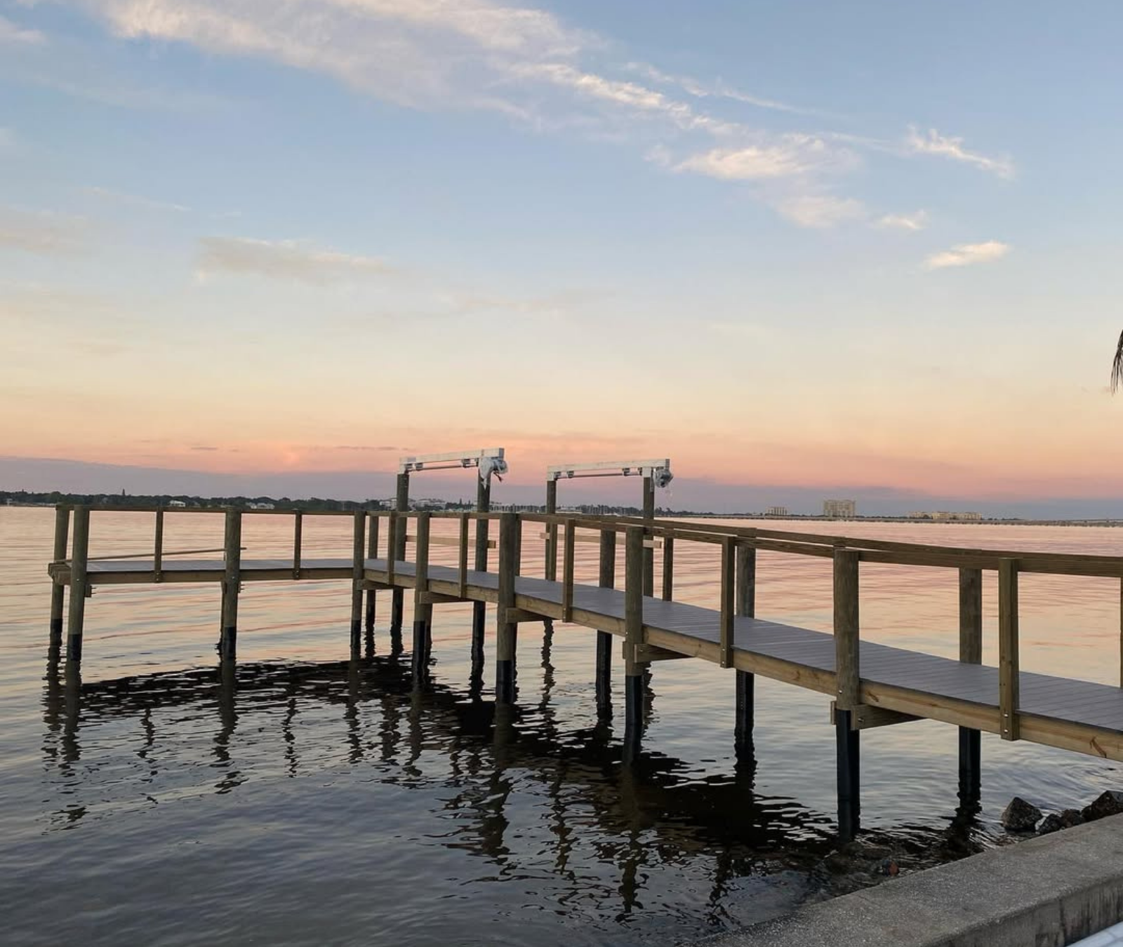 A wooden dock extends over calm water at sunset, with soft pink and blue hues in the sky.