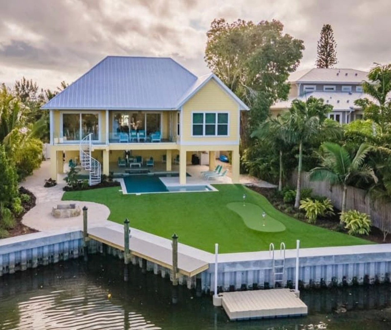A yellow waterfront house with a metal roof, swimming pool, green lawn, putting green, and a private dock on a canal.