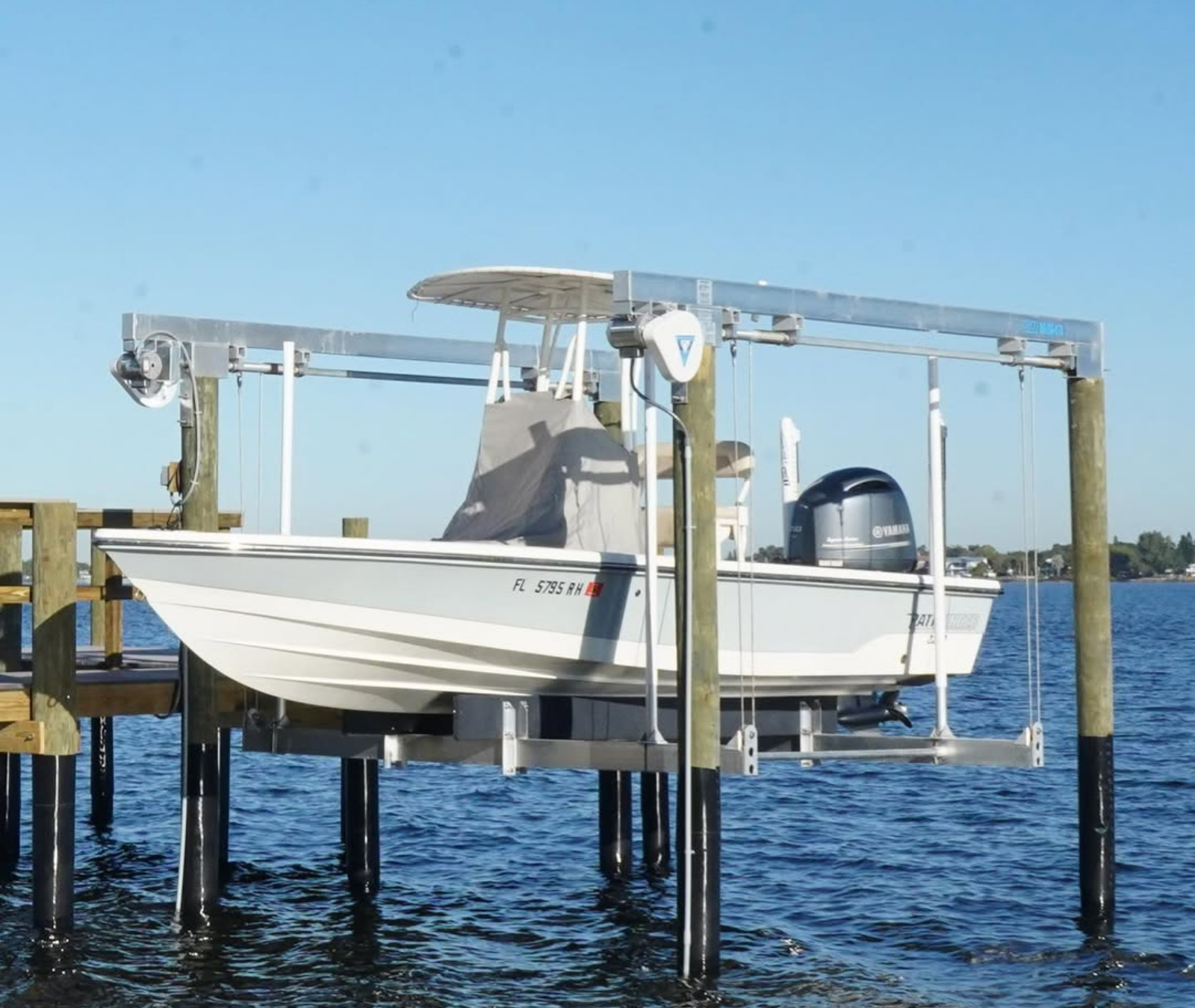 A light-colored motorboat with a gray center console sits raised on a mechanical boat lift above blue water.