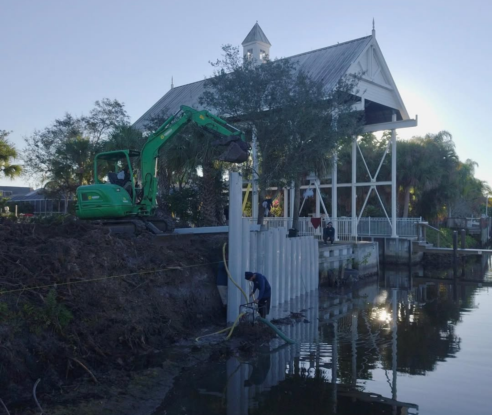 A green excavator installs white vinyl seawall panels along a canal bank next to a white structure with a cupola.