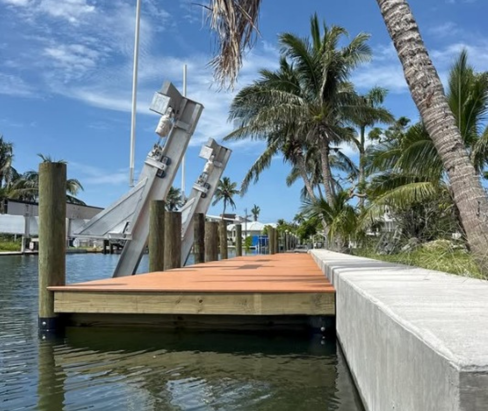 A new wooden dock with metal boat lift posts extends over calm canal water next to a concrete seawall and palm trees.