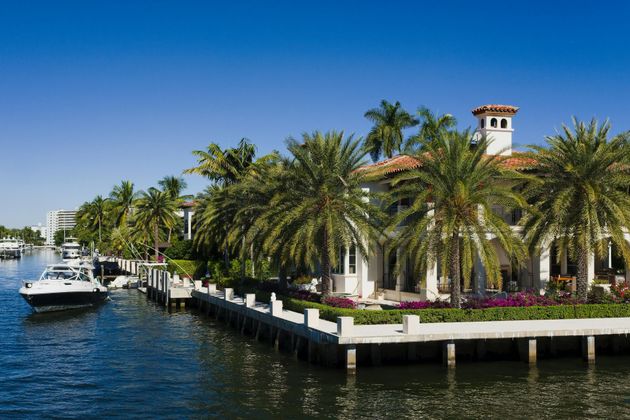 Luxury waterfront marina with yachts, palm trees, and a Mediterranean-style building under a clear blue sky