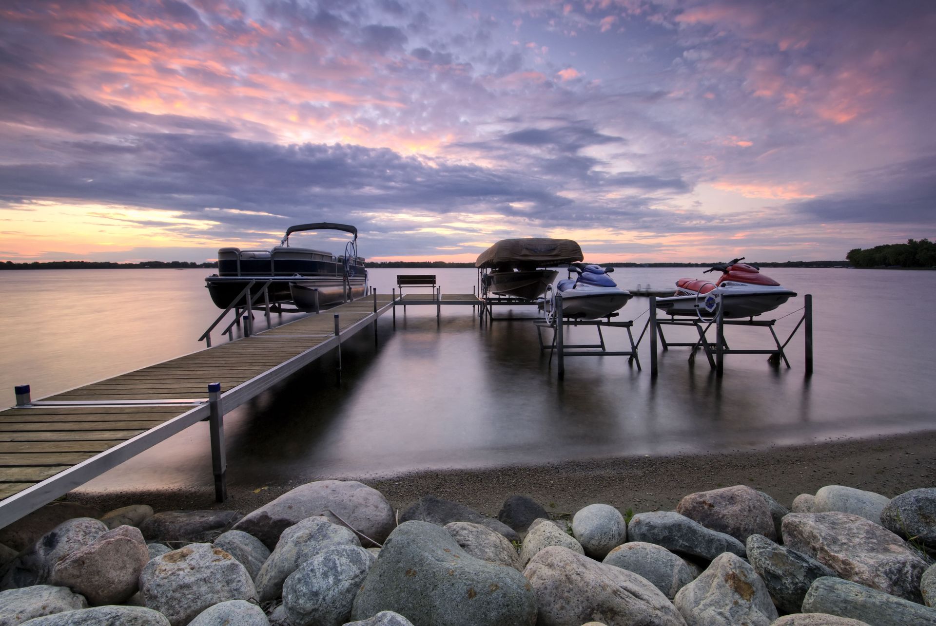 Dock with lounge chairs and pontoon boats on calm water at sunset, with rocks in the foreground.