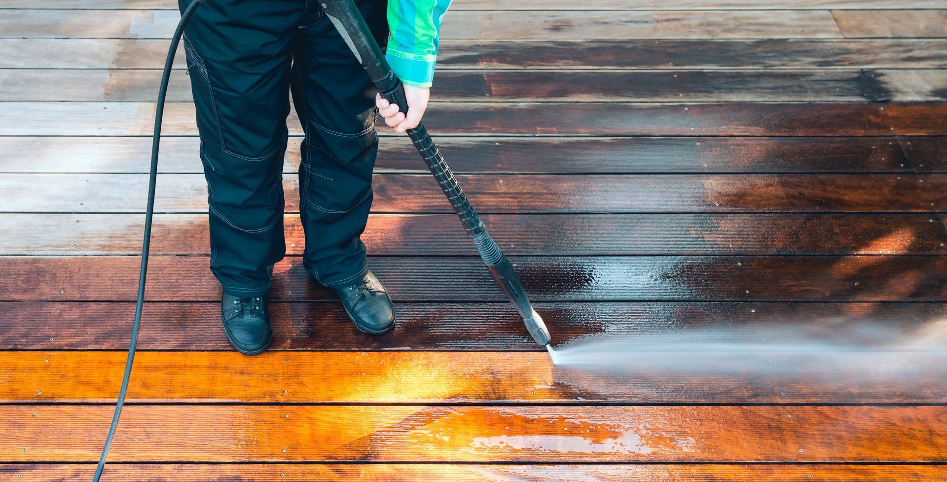 A person in dark work clothes pressure washes a weathered wooden deck, revealing a clean, bright, orange-toned surface.