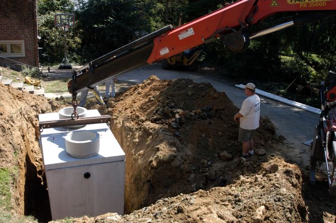 Excavator installing a large concrete tank in a dirt excavation beside a worker outdoors.