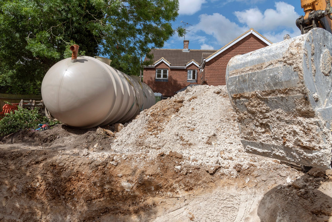 Large white cylindrical tank lying on a dirt mound near houses and trees under a blue sky