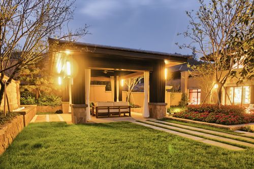 Warmly lit modern house with a covered patio, green lawn, and garden at dusk