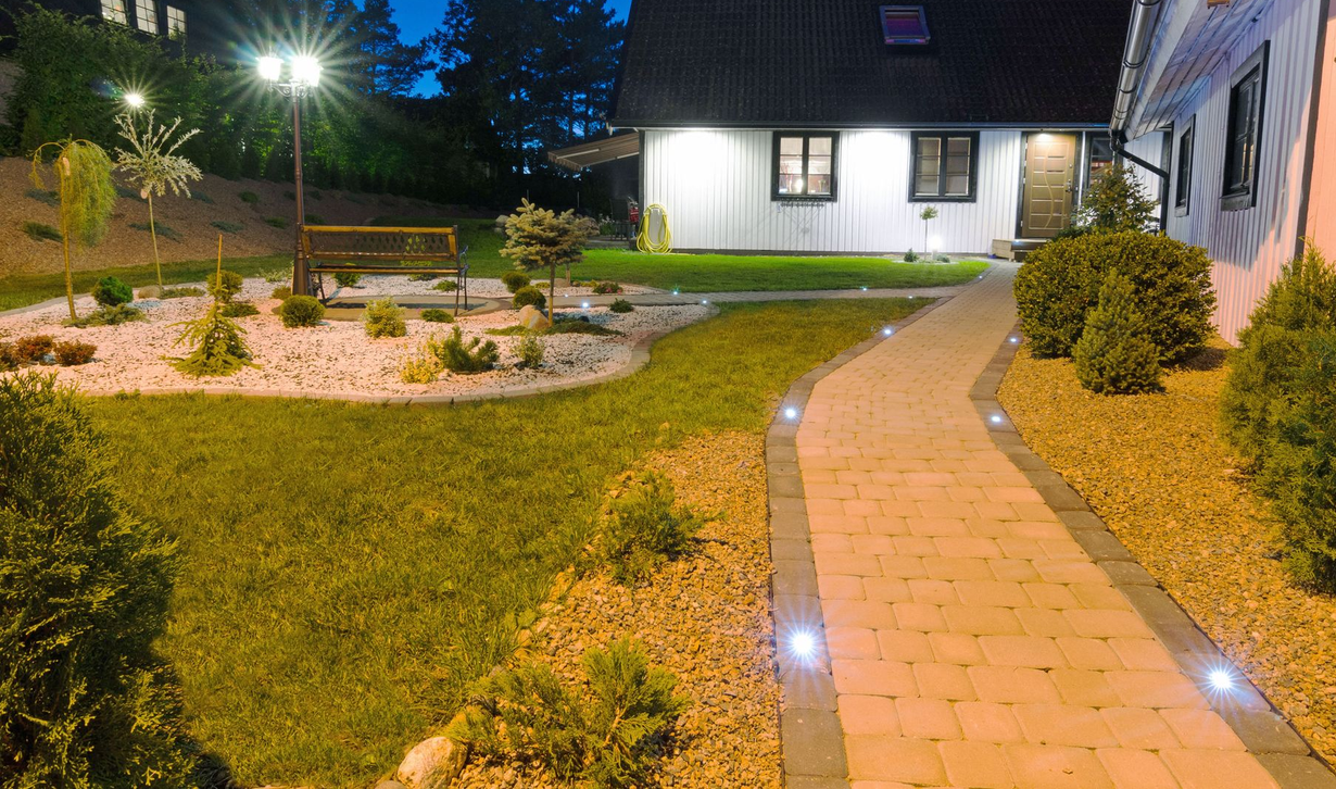Lit garden path beside a house at dusk, with lawn, shrubs, and landscape lights.
