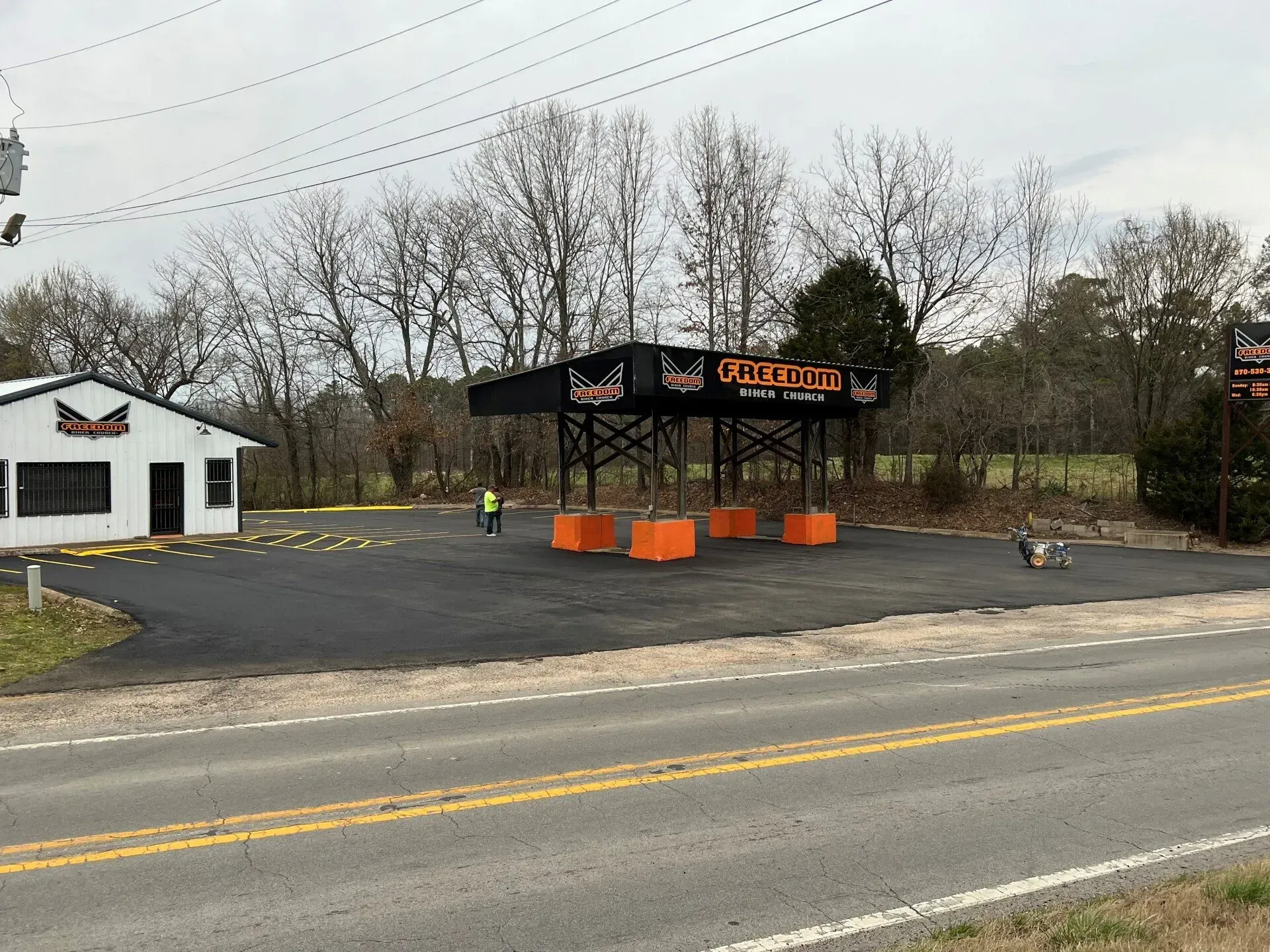 Small roadside food stand with black canopy and orange barriers beside a road, trees in the background