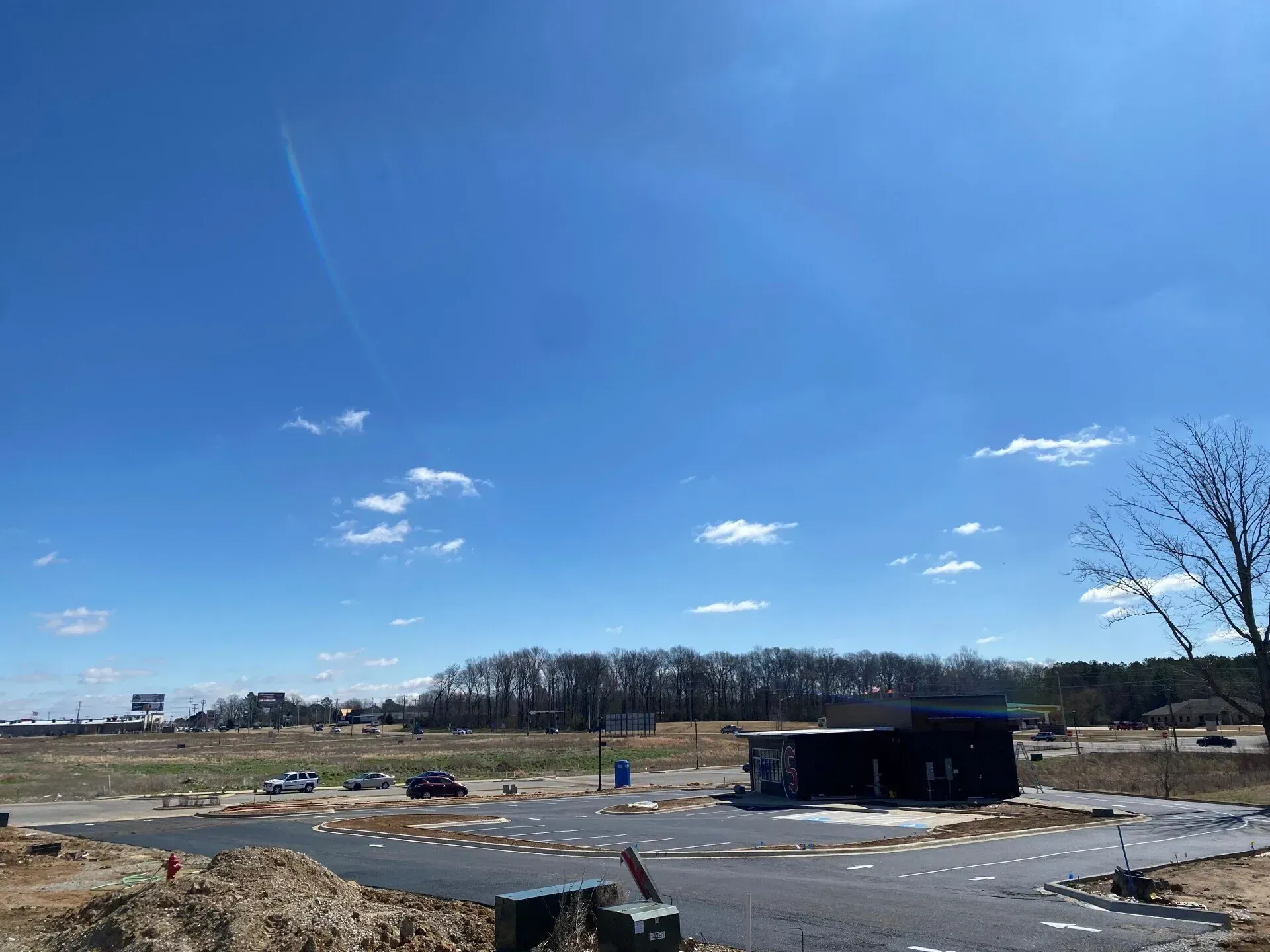 Open field with a small dark building, cars, and snow patches under a bright blue sky