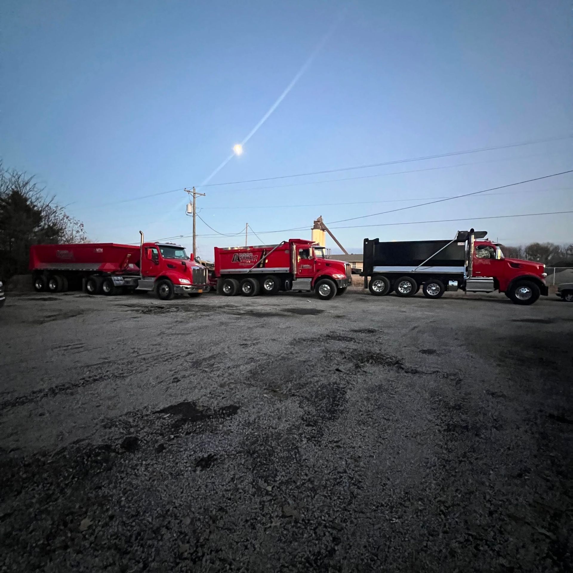 Four red dump trucks and one black dump truck parked in a gravel lot at dusk
