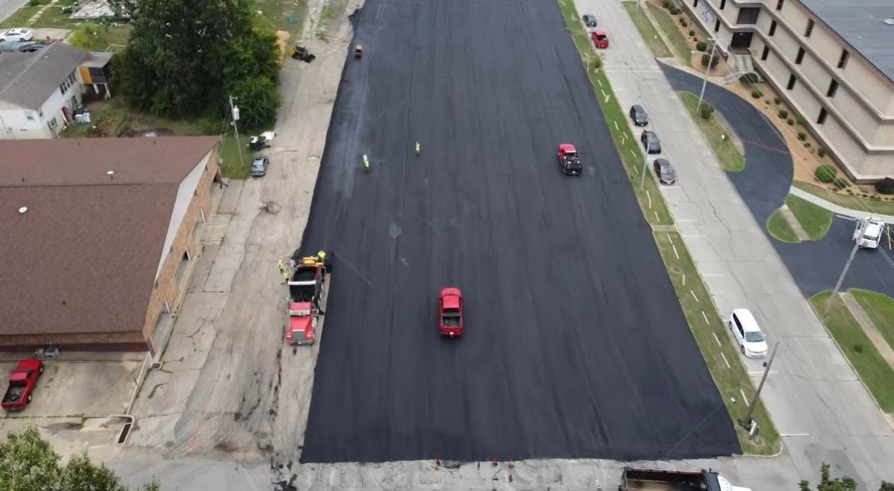 Aerial view of fresh black asphalt paving with several red work vehicles on a city street.