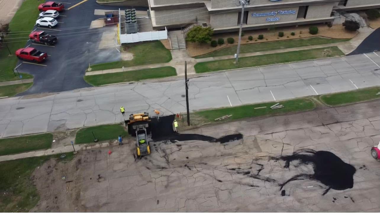 Aerial view of road construction with excavator on a dirt lot beside a street and parked cars
