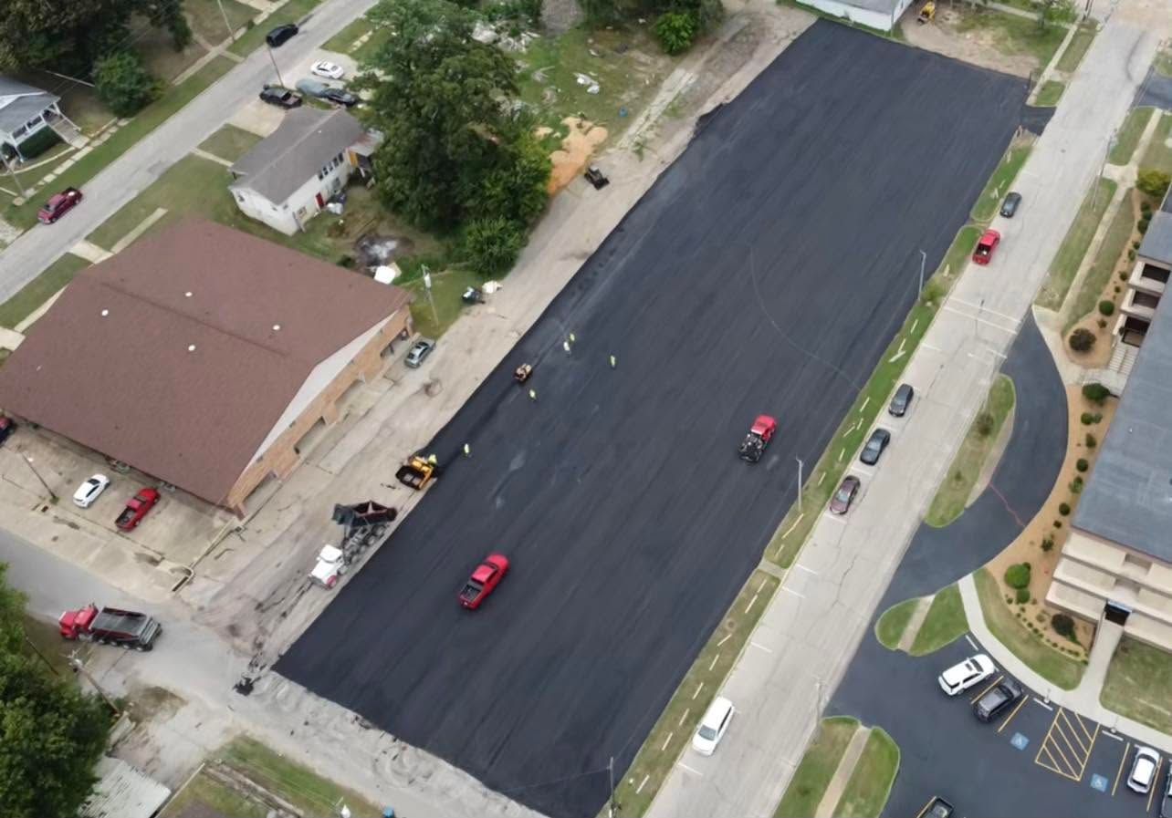 Aerial view of a freshly paved black parking lot beside buildings, with a few cars and workers nearby